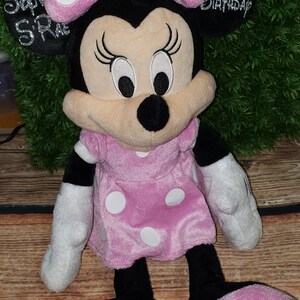Może przedstawiać: Pluszowa zabawka Minnie Mouse w kolorze r&oacute;żowym i czarnym z białymi kropkami. Ma na sobie r&oacute;żową sukienkę w białe kropki i r&oacute;żową kokardkę w białe kropki. Zabawka ma czarny nos i haftowane oczy. Tekst "1. urodziny" jest wyszyty na tylnej części głowy zabawki.