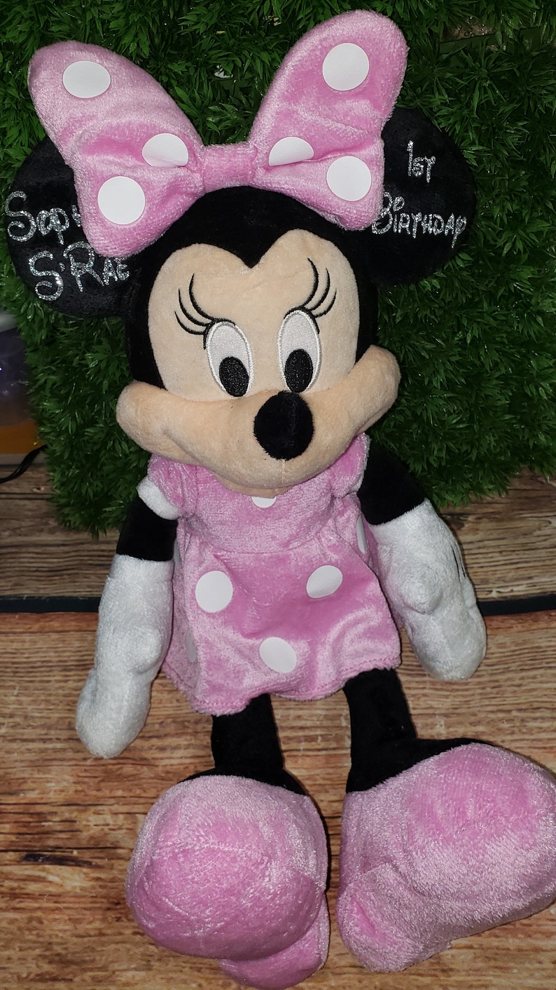 Może przedstawiać: Pluszowa zabawka Minnie Mouse w kolorze r&oacute;żowym i czarnym z białymi kropkami. Ma na sobie r&oacute;żową sukienkę w białe kropki i r&oacute;żową kokardkę w białe kropki. Zabawka ma czarny nos i haftowane oczy. Tekst "1. urodziny" jest wyszyty na tylnej części głowy zabawki.