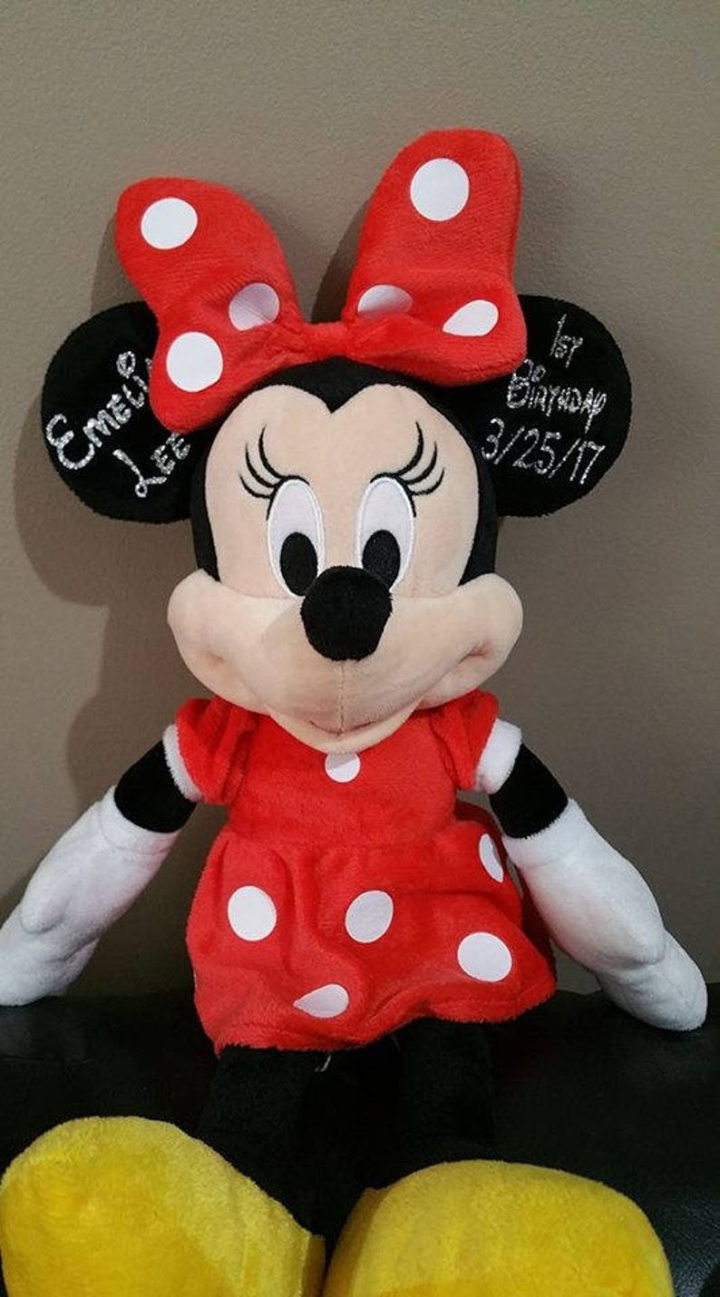 Może przedstawiać: Pluszowa zabawka Minnie Mouse w czerwono-białe kropki z czarnymi uszami i czarnym nosem. Zabawka ma na sobie czerwoną sukienkę w białe kropki i ż&oacute;łte buty. Zabawka ma wyszyty tekst, kt&oacute;ry brzmi "Emilee Lee 1. urodziny 3/25/17".