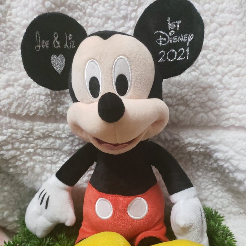 Może przedstawiać: Pluszowa zabawka Mickey Mouse w kolorze czarnym i białym, w czerwonych spodenkach i ż&oacute;łtych butach. Uszy są wyszyte napisem "Joe & Liz" i sercem na jednym uchu oraz "1st Disney 2021" na drugim.