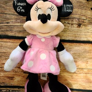 Może przedstawiać: R&oacute;żowa pluszowa zabawka Minnie Mouse w białe kropki, w r&oacute;żowej kokardce z białymi kropkami. Zabawka ma wyszyty tekst na uchu, kt&oacute;ry brzmi "Flower Girl 7/1/2020".