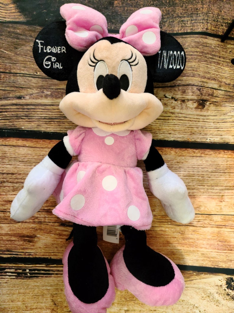 Może przedstawiać: R&oacute;żowa pluszowa zabawka Minnie Mouse w białe kropki, w r&oacute;żowej kokardce z białymi kropkami. Zabawka ma wyszyty tekst na uchu, kt&oacute;ry brzmi "Flower Girl 7/1/2020".