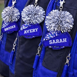 May include: Three blue and black backpacks with personalized cheerleader-themed keychains. Each keychain features a silver pom-pom and a blue megaphone with a name: Brooke, Avery, and Sarah. The backpacks have blue accents.