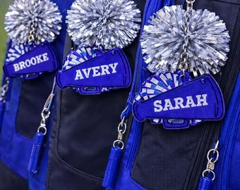 Peut inclure: Trois sacs à dos bleus et noirs avec des porte-clés personnalisés sur le thème des cheerleaders. Chaque porte-clés comprend un pompon argenté et un mégaphone bleu avec un nom : Brooke, Avery et Sarah. Les sacs à dos ont des accents bleus.
