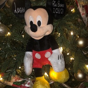 Może przedstawiać: Pluszowa zabawka Mickey Mouse w kolorze czarnym i czerwonym z ż&oacute;łtymi butami wisi na choince. Zabawka ma metkę z imieniem "Logan" i metkę z napisem "Ring Bearer 2/28/21".