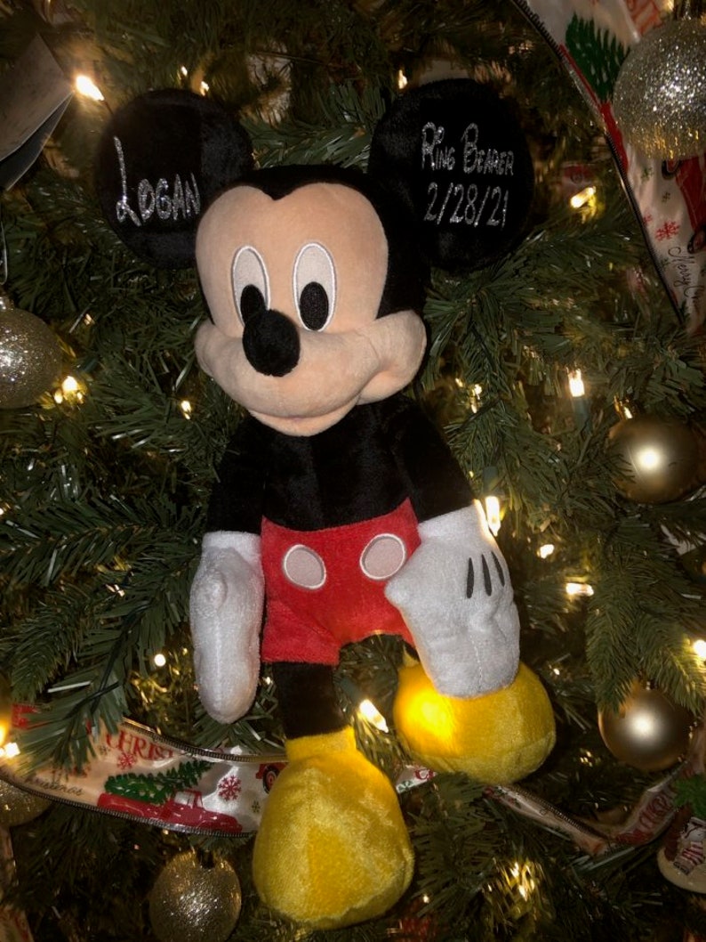 Może przedstawiać: Pluszowa zabawka Mickey Mouse w kolorze czarnym i czerwonym z ż&oacute;łtymi butami wisi na choince. Zabawka ma metkę z imieniem "Logan" i metkę z napisem "Ring Bearer 2/28/21".