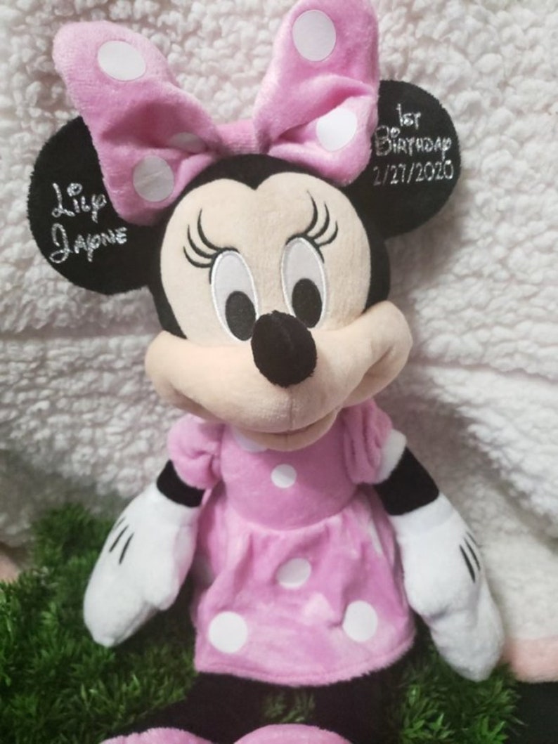 Może przedstawiać: Pluszowa myszka Minnie w r&oacute;żowo-białe kropki z czarną kokardką i haftowanymi oczami. Zabawka ma wyszyte napisy "Lily Jayne" i "1. urodziny 2/21/2020".