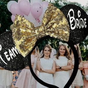 May include: Black headband with gold glitter bow and two black ears. One ear says "Team" and the other says "Bride".