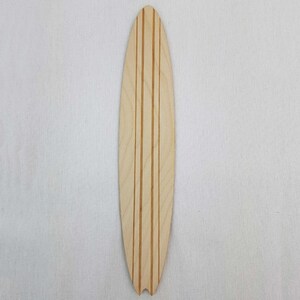 May include: A wooden surfboard cutout with three parallel lines carved into the surface. The surfboard is light brown and has a rounded shape.