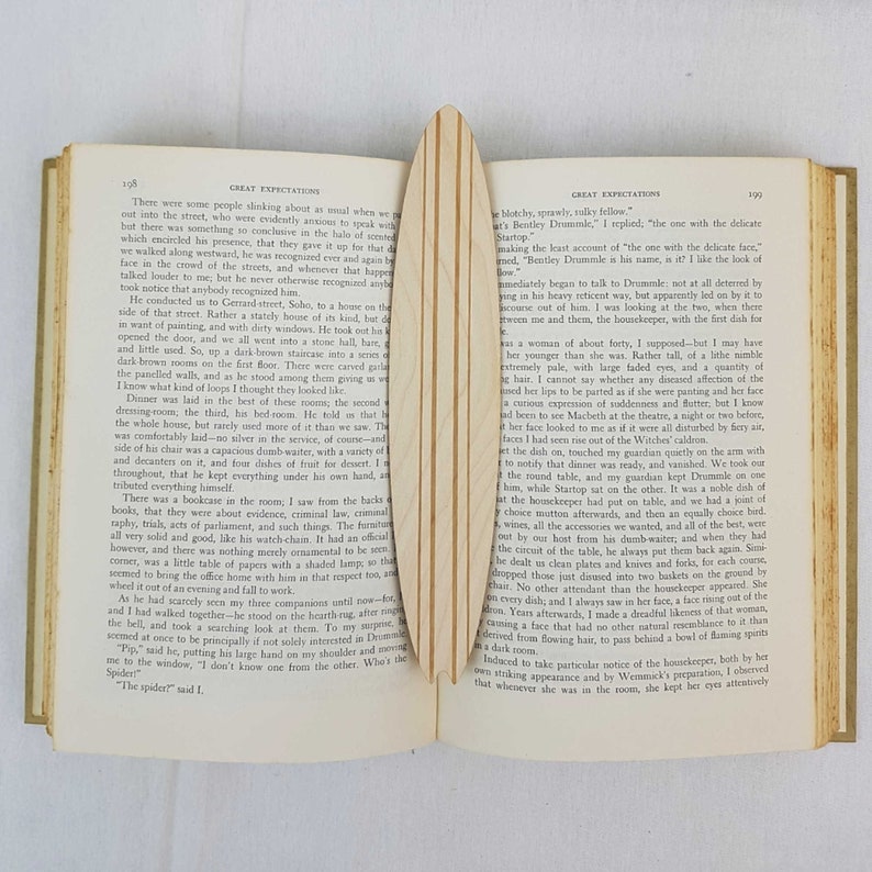 May include: An open book with a wooden bookmark. The book is titled "Great Expectations" and the pages are filled with text. The bookmark is a light brown color and has a smooth, polished surface.