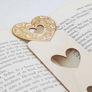 May include: Two wooden heart-shaped bookmarks with a cutout design. The bookmark on the left has a gold floral pattern. The bookmark on the right is plain wood. The bookmarks are placed on a book with text.