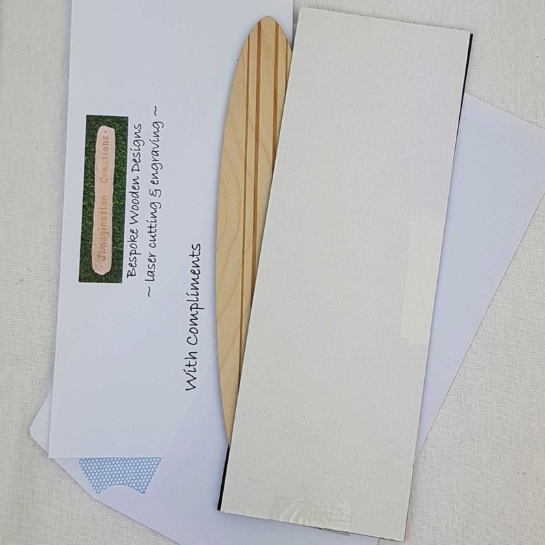 May include: A wooden surfboard bookmark with a natural wood finish and three brown stripes. The bookmark is on a white background with the text "With Compliments" and "Bespoke Wooden Designs - laser cutting & engraving - Imagination Creations".