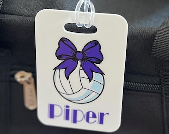 May include: A black duffel bag with a white tag attached. The tag features a volleyball design with a purple bow and the name "Piper" in purple text. The bag has a zipper and black straps.