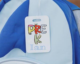 May include: A rectangular white plastic luggage tag attached to a blue shark-themed backpack. The tag features the colorful text PRE K with a small apple icon above the K, and the name Ian printed in light blue script font below the text.