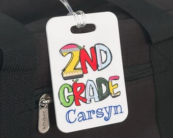 May include: A white rectangular luggage tag attached to a black duffel bag. The tag features the text 2ND GRADE in colorful, patterned block letters with a small red apple icon, and the name Carsyn printed in blue script below the text.