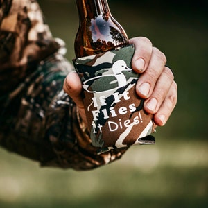 May include: A camouflage-patterned can cooler with the text "If It Flies It Dies!" and a white silhouette of a duck. The cooler is holding a brown glass bottle.