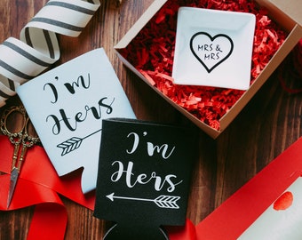 May include: A collection of wedding-themed gifts and accessories. Includes two can coolers with "I'm Hers" text, a white dish with "MRS & MRS" in a heart, and a gift box with red paper filler. Decorative ribbons and wrapping paper are also present.