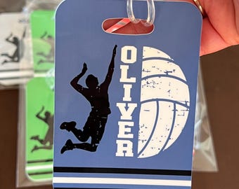May include: Blue luggage tag with a volleyball design. A black silhouette of a volleyball player jumping to spike the ball is featured. The name "OLIVER" is printed vertically. The tag has a clear plastic loop for attachment.