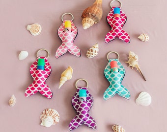 May include: Six colorful mermaid tail-shaped keychains with different colored lip balm tubes inside. The keychains are on a pink background with seashells scattered around.