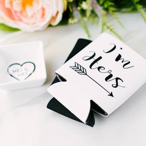 May include: White and black drink koozies with the text "I'm Hers" and an arrow. A small white square dish with a heart and the words "Mrs & Mrs" is also present. The items are arranged on a white surface with floral accents.