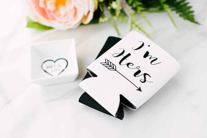 May include: White and black drink koozies with the text "I'm Hers" and an arrow. A small white square dish with a heart and the words "Mrs & Mrs" is also present. The items are arranged on a white surface with floral accents.