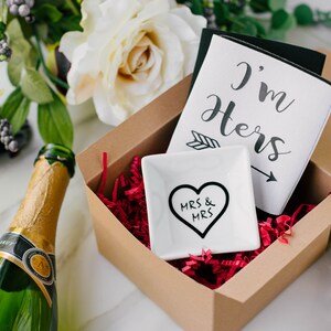 May include: A gift set in a brown box with red filler, including a white dish with a black heart and "MRS & MRS" text, and a card that says "I'm Hers". A green champagne bottle is visible.