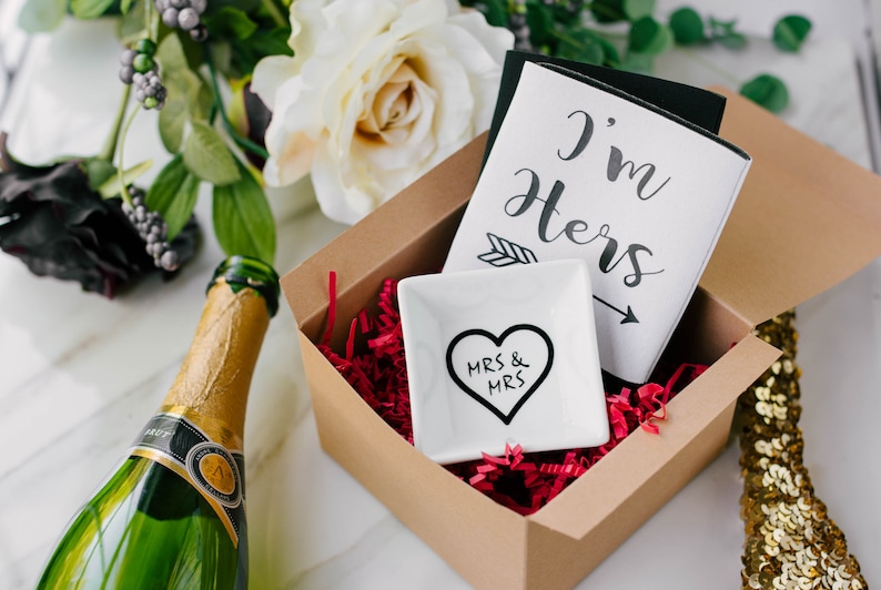 May include: A gift set in a brown box with red filler, including a white dish with a black heart and "MRS & MRS" text, and a card that says "I'm Hers". A green champagne bottle is visible.