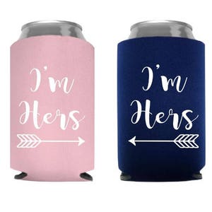 May include: Two can coolers, one pink and one navy blue, each with the text "I'm Hers" in white script, accompanied by a white arrow graphic. The coolers are designed to keep beverages cold.