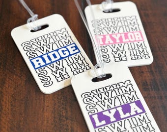 May include: Three white luggage tags with clear plastic loops. Each tag has the word "SWIM" repeated in a pattern. One tag has "RIDGE" in blue, another has "TAYLOR" in pink, and the third has "LYLA" in purple.