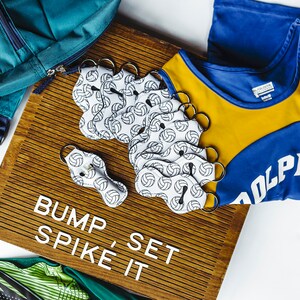 May include: A set of 10 white volleyball keychain lip balms with a black volleyball pattern. The lip balms are arranged on a wooden board with the words "Bump, Spike, Set It" written on it. A blue and yellow volleyball jersey with the word "Dolphin" on it is in the background.