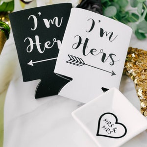 May include: Black and white beverage insulators with the text "I'm Her" and "I'm Hers" with an arrow. A small white square dish with a heart that says "Mrs & Mrs" is also in the image. A bottle of champagne and a gold sequined hanger are also visible.