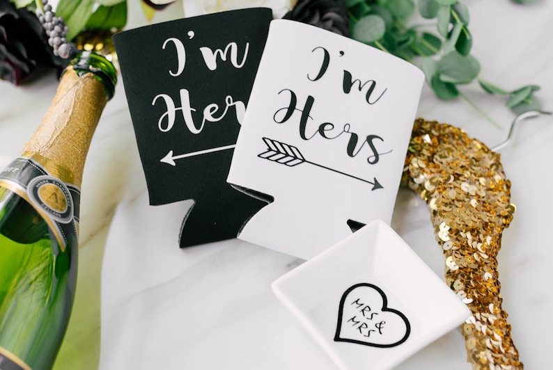 May include: Black and white beverage insulators with the text "I'm Her" and "I'm Hers" with an arrow. A small white square dish with a heart that says "Mrs & Mrs" is also in the image. A bottle of champagne and a gold sequined hanger are also visible.