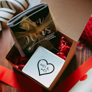 May include: A gift box with a black notebook tied with a gold bow, a white dish with a heart design, and red paper filler. The notebook has the words "Mrs & Mrs" and an arrow. The box is on a wooden surface with red ribbon and wrapping paper.