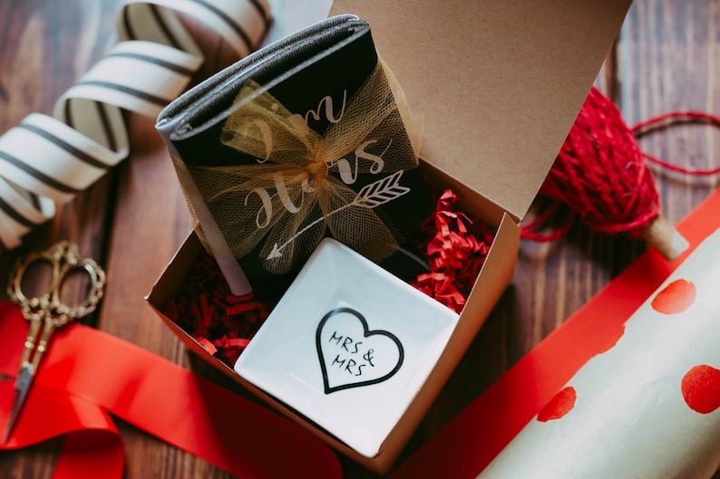 May include: A gift box with a black notebook tied with a gold bow, a white dish with a heart design, and red paper filler. The notebook has the words "Mrs & Mrs" and an arrow. The box is on a wooden surface with red ribbon and wrapping paper.