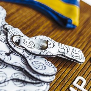 May include: A white keychain with a volleyball pattern and black trim. The keychain has a silver ring and is on a wooden surface with the word "SET" in white letters.