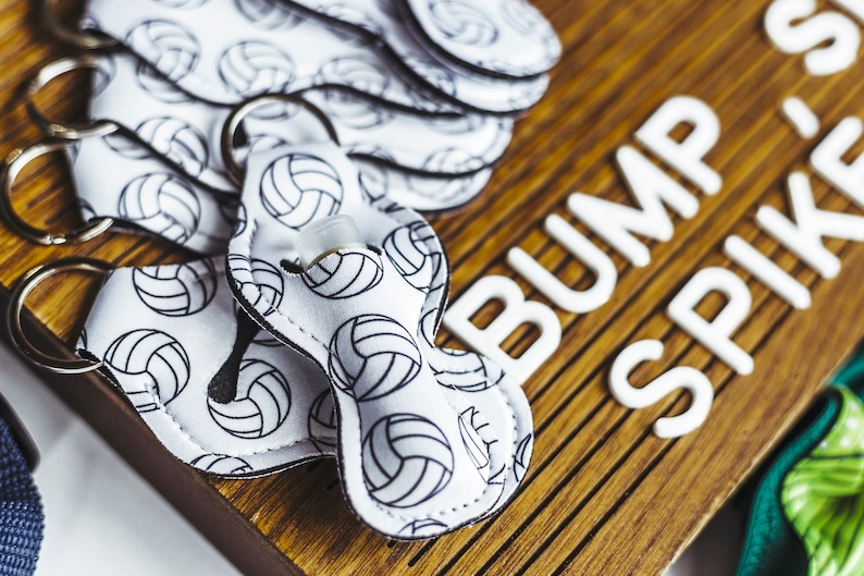 May include: White keyrings with a volleyball pattern and black trim. The keyrings have a small clear bottle attached to them. The keyrings are on a wooden surface with the words "BUMP SET SPIKE" written in white letters.