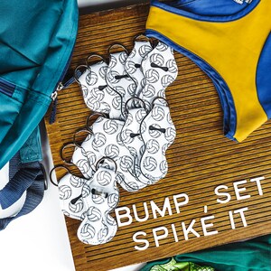 May include: A wooden sign with the text "Bump, Set, Spike It" and a volleyball-themed keychain with white and black volleyballs on a white background. The sign is surrounded by a blue, yellow, and green volleyball uniform.