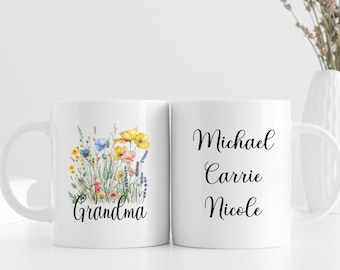 May include: Two white ceramic mugs. One mug features a watercolor floral design with the word "Grandma". The other mug has the names "Michael", "Carrie", and "Nicole" in elegant script. A vase with dried flowers is in the background.