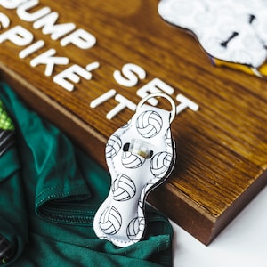 May include: White fabric keychain with a volleyball pattern and a silver ring. The keychain is on a wooden surface with the words "Bump Spike It Set" written in white letters.