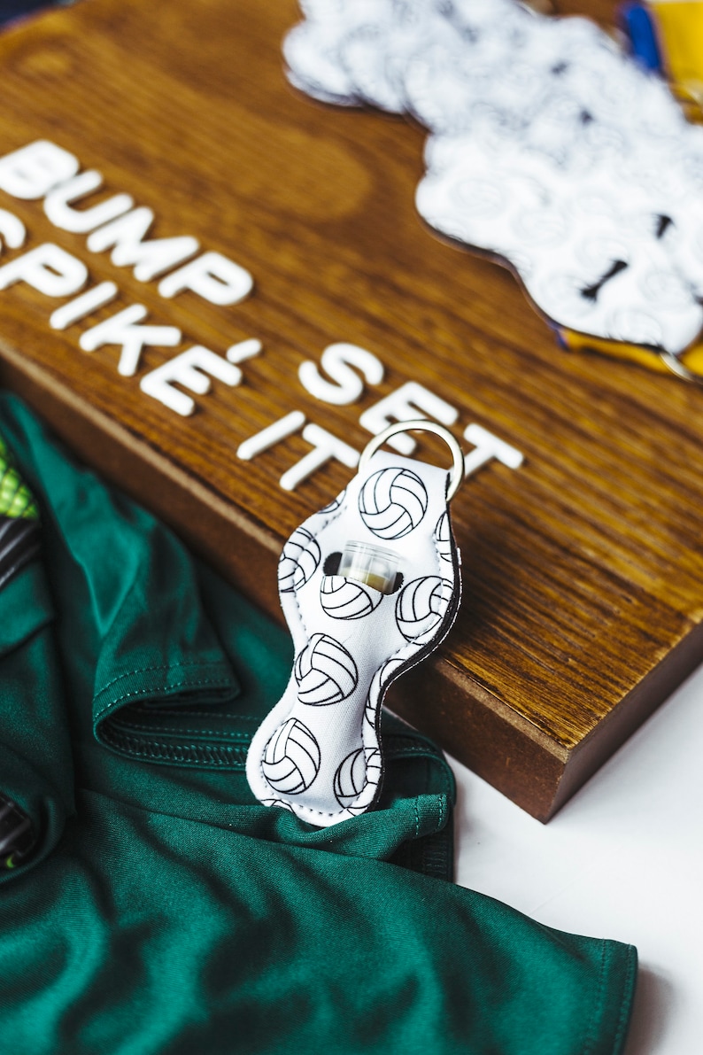 May include: White fabric keychain with a volleyball pattern and a silver ring. The keychain is on a wooden surface with the words "Bump Spike It Set" written in white letters.