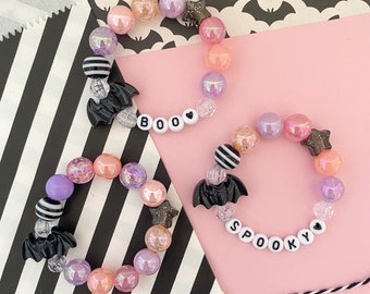 Könnte beinhalten: Zwei bunte Perlenarmbänder mit schwarzen Fledermaus-Anhängern und Buchstabenperlen, die "BOO" und "SPOOKY" buchstabieren.