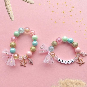 May include: Two beaded bracelets with pink and blue beads, gold glitter, and a starfish charm. One bracelet has a pink tulle bow and the other has a white tulle bow. The bracelet with the tulle bow has the name "ARIEL" spelled out in white beads.