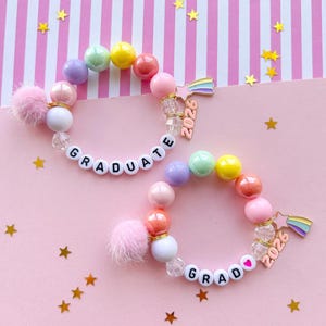 May include: Two beaded bracelets with colorful beads, white letter beads spelling "GRADUATE", and a charm that says "2026". One bracelet has a pink pom-pom. The background is pink with gold star confetti.
