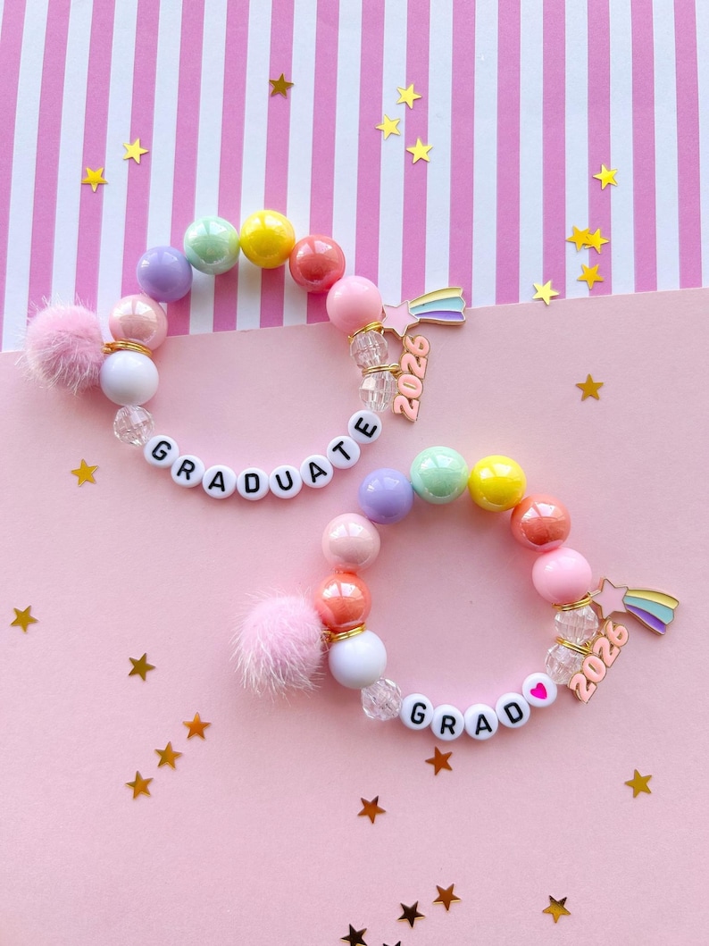May include: Two beaded bracelets with colorful beads, white letter beads spelling "GRADUATE", and a charm that says "2026". One bracelet has a pink pom-pom. The background is pink with gold star confetti.