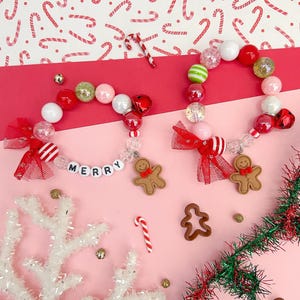 May include: Two Christmas-themed beaded bracelets with red, white, and green beads, a gingerbread man charm, and the word "MERRY" spelled out in white beads. The bracelets are on a pink surface with red and white candy canes, a white snowflake, and green and red tinsel.