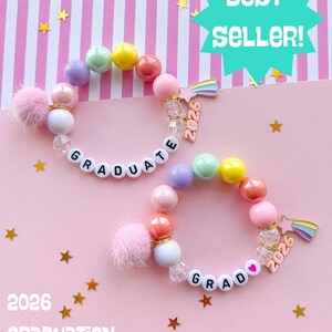 May include: Two beaded bracelets with the word "GRADUATE" spelled out in white letter beads. Each bracelet features a variety of colorful beads, a pink pom-pom, and a charm with the year 2026. The background is pink with gold stars.