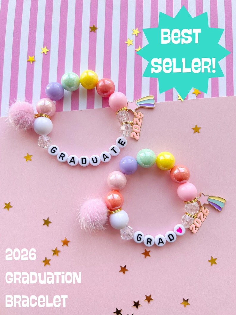 May include: Two beaded bracelets with the word "GRADUATE" spelled out in white letter beads. Each bracelet features a variety of colorful beads, a pink pom-pom, and a charm with the year 2026. The background is pink with gold stars.