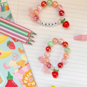 May include: Two beaded bracelets with the name "LOGAN" spelled out in white letter beads. The bracelets have a variety of colorful beads, a red apple charm, and a pink bow. Pencils and a snack-themed notepad are also visible.