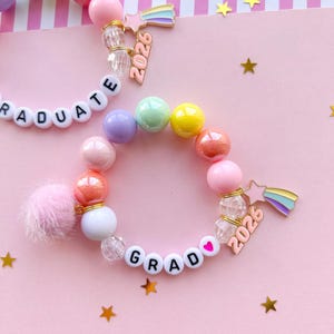 May include: Two beaded bracelets with colorful round beads, white letter beads spelling "GRAD" and "GRADUATE", and a pink pom-pom. Each bracelet has a charm with a rainbow and the number "2026". The background is pink with gold stars.