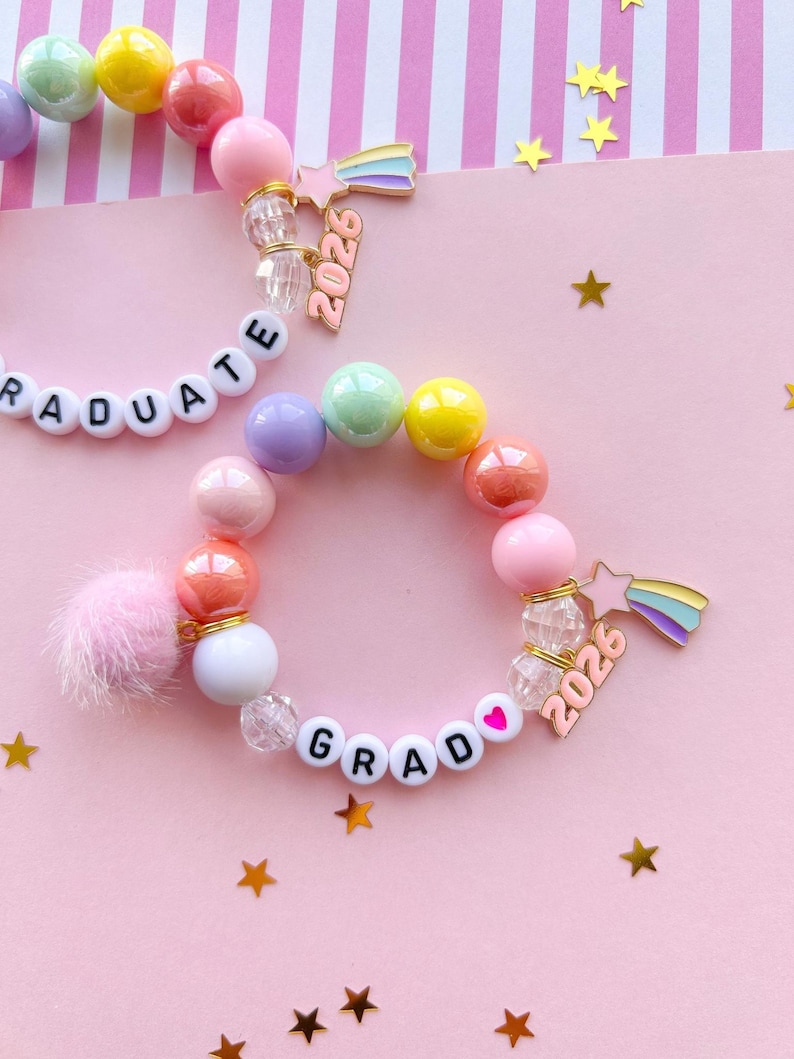 May include: Two beaded bracelets with colorful round beads, white letter beads spelling "GRAD" and "GRADUATE", and a pink pom-pom. Each bracelet has a charm with a rainbow and the number "2026". The background is pink with gold stars.